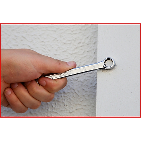 Eine Hand hält einen silbernen Ringschlüssel, der an eine weiße Wand gehalten wird. Der Hintergrund ist strukturiert.